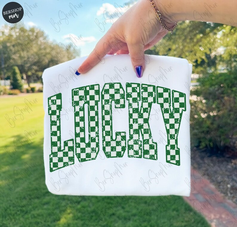 May include: White sweatshirt with the word "LUCKY" in large, green, checkered letters. The sweatshirt is held up against a background of green foliage and a blue sky. The design is a bold, eye-catching statement.