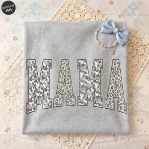 May include: A light gray sweatshirt with the word "MAMA" printed across the front. The letters have a camouflage and leopard print design. A light blue bow and pearl bracelet are on the top right corner.