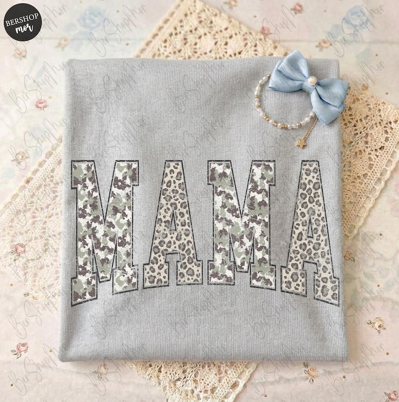May include: A light gray sweatshirt with the word "MAMA" printed across the front. The letters have a camouflage and leopard print design. A light blue bow and pearl bracelet are on the top right corner.