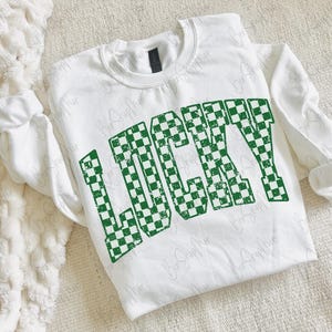 May include: White sweatshirt with the word "LUCKY" in large, green, checkered letters. The sweatshirt has long sleeves and a crew neck. The design is slightly distressed, giving it a vintage look. The sweatshirt is likely made of a soft, comfortable material.