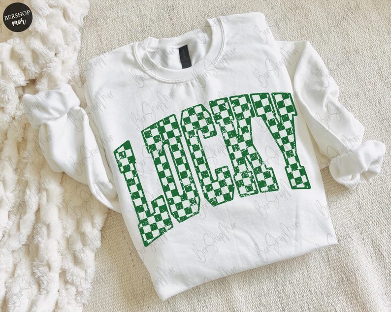 May include: White sweatshirt with the word "LUCKY" in large, green, checkered letters. The sweatshirt has long sleeves and a crew neck. The design is slightly distressed, giving it a vintage look. The sweatshirt is likely made of a soft, comfortable material.