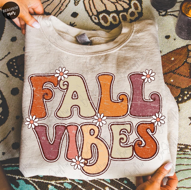 May include: A beige sweatshirt with the words "FALL VIBES" in retro-style lettering. The letters are in shades of orange, beige, and burgundy, with small daisy accents. The sweatshirt is folded and held by hands.