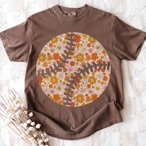 May include: Brown t-shirt featuring a floral baseball design. The baseball graphic is a circle filled with orange, yellow, and pink flowers, with brown stitching details. The shirt has short sleeves and a crew neck.