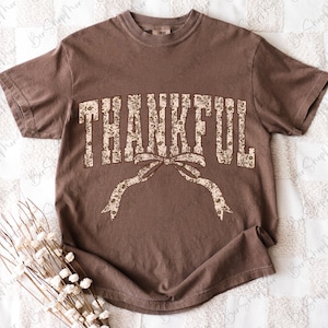 May include: A brown t-shirt with the word "THANKFUL" in a floral design across the chest. The word is arched with a bow underneath. The shirt has short sleeves and a crew neck.