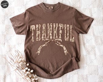 May include: A brown t-shirt with the word "THANKFUL" in a floral design across the chest. The word is arched with a bow underneath. The shirt has short sleeves and a crew neck.