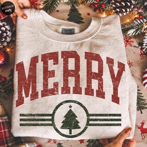 Puede incluir: Una sudadera de cuello redondo color crema con la palabra MERRY en letras mayúsculas rojas desgastadas. Debajo del texto hay un emblema circular verde oscuro que contiene un árbol de Navidad estilizado, flanqueado por tres líneas horizontales.