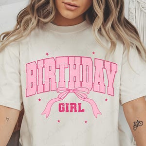 May include: Cream-colored t-shirt with the words "BIRTHDAY GIRL" in pink block letters. A pink bow and ribbon design is below the text, with pink star accents. The shirt has short sleeves.
