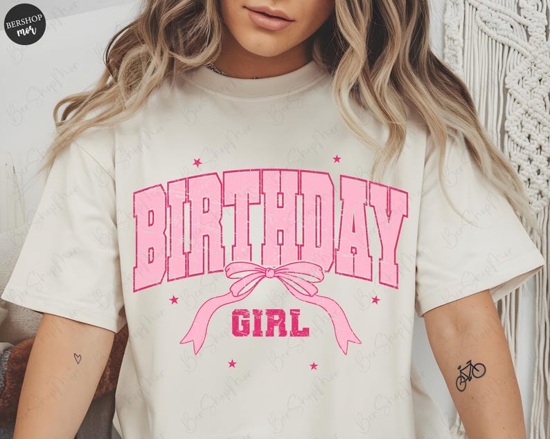 May include: Cream-colored t-shirt with the words "BIRTHDAY GIRL" in pink block letters. A pink bow and ribbon design is below the text, with pink star accents. The shirt has short sleeves.