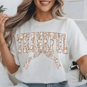 May include: White t-shirt with the word "THANKFUL" in a floral design. A decorative bow with a floral pattern is below the word. The shirt has short sleeves and a crew neck.