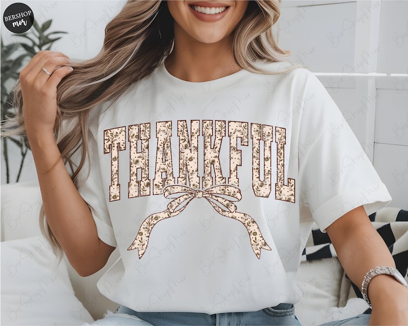 May include: White t-shirt with the word "THANKFUL" in a floral design. A decorative bow with a floral pattern is below the word. The shirt has short sleeves and a crew neck.