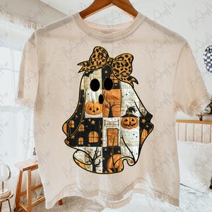 May include: Cream-colored t-shirt featuring a Halloween ghost design. The ghost is a patchwork of black, orange, and white squares, with a leopard print bow. The design includes jack-o'-lanterns, houses, and trees.