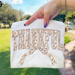 May include: A white sweatshirt with the word "THANKFUL" in a brown floral design. A brown floral bow is below the word. The sweatshirt is held up by a person.