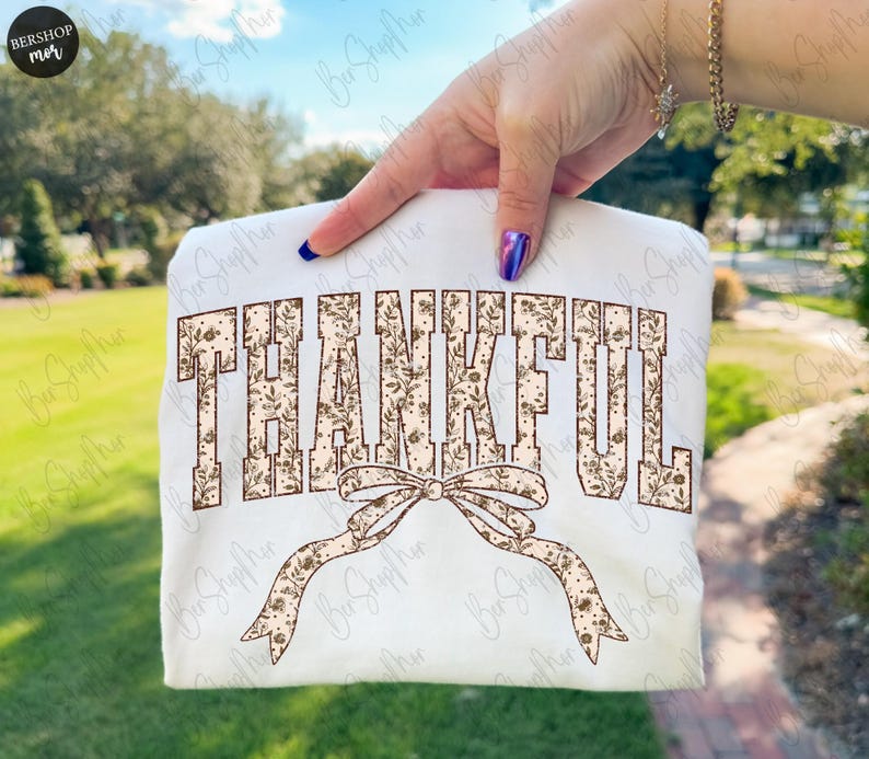 May include: A white sweatshirt with the word "THANKFUL" in a brown floral design. A brown floral bow is below the word. The sweatshirt is held up by a person.