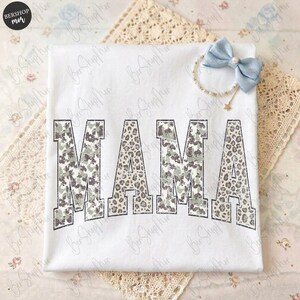 May include: White t-shirt with the word "MAMA" in large, bold letters. The letters have a camouflage and leopard print design. A light blue bow and bracelet with a star charm are also visible.