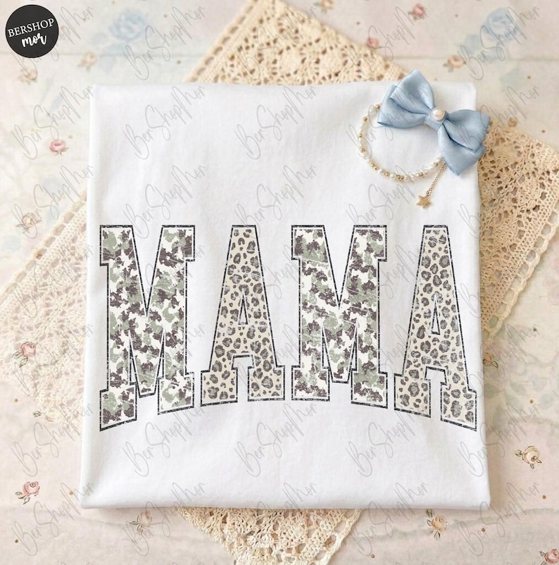 May include: White t-shirt with the word "MAMA" in large, bold letters. The letters have a camouflage and leopard print design. A light blue bow and bracelet with a star charm are also visible.