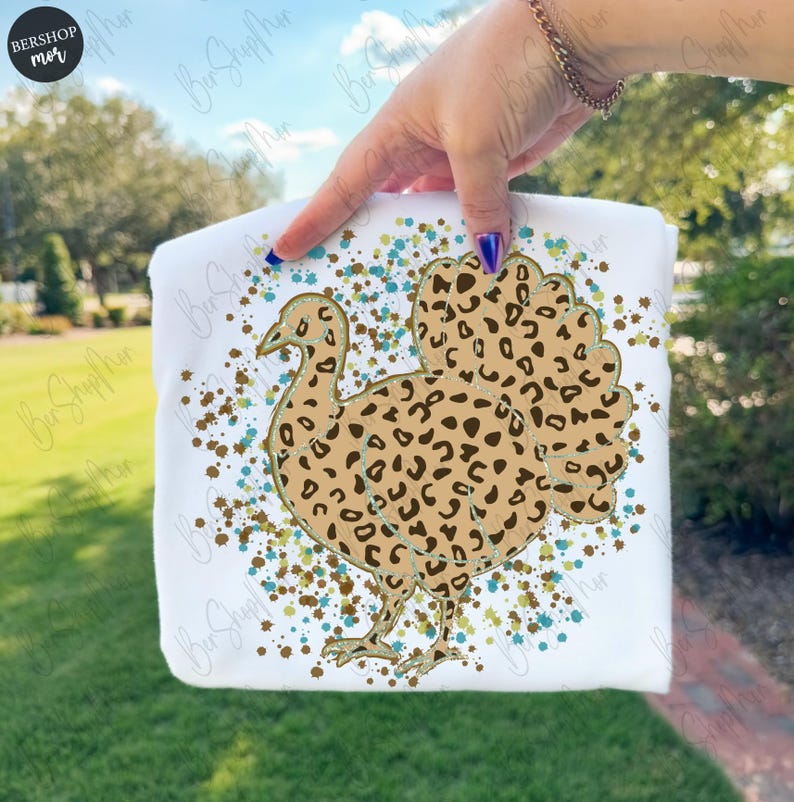 May include: White t-shirt featuring a turkey design with a leopard print pattern. The turkey is outlined in a light brown color and is surrounded by blue and brown paint splatters. A fun and festive design.