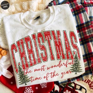 Op de afbeelding: Een wit sweatshirt met ronde hals voorzien van de rode, verweerde tekst CHRISTMAS in een groot collegiaal lettertype, gevolgd door de rode cursieve tekst the most wonderful time of the year, geaccentueerd door twee groene dennenbomen.