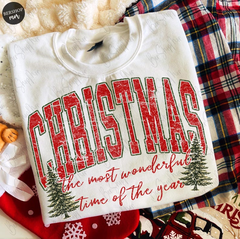 Op de afbeelding: Een wit sweatshirt met ronde hals voorzien van de rode, verweerde tekst CHRISTMAS in een groot collegiaal lettertype, gevolgd door de rode cursieve tekst the most wonderful time of the year, geaccentueerd door twee groene dennenbomen.