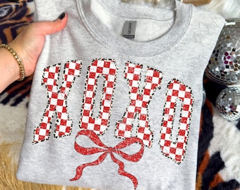 May include: A light gray sweatshirt with the letters "XOXO" in a red and white checkered pattern. A red bow is below the letters. The sweatshirt is a Gildan brand.
