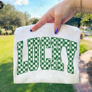 May include: A white t-shirt with the word "LUCKY" printed in large, green, checkered letters. The text has a distressed, vintage look. The shirt is held up against a blurred outdoor background.