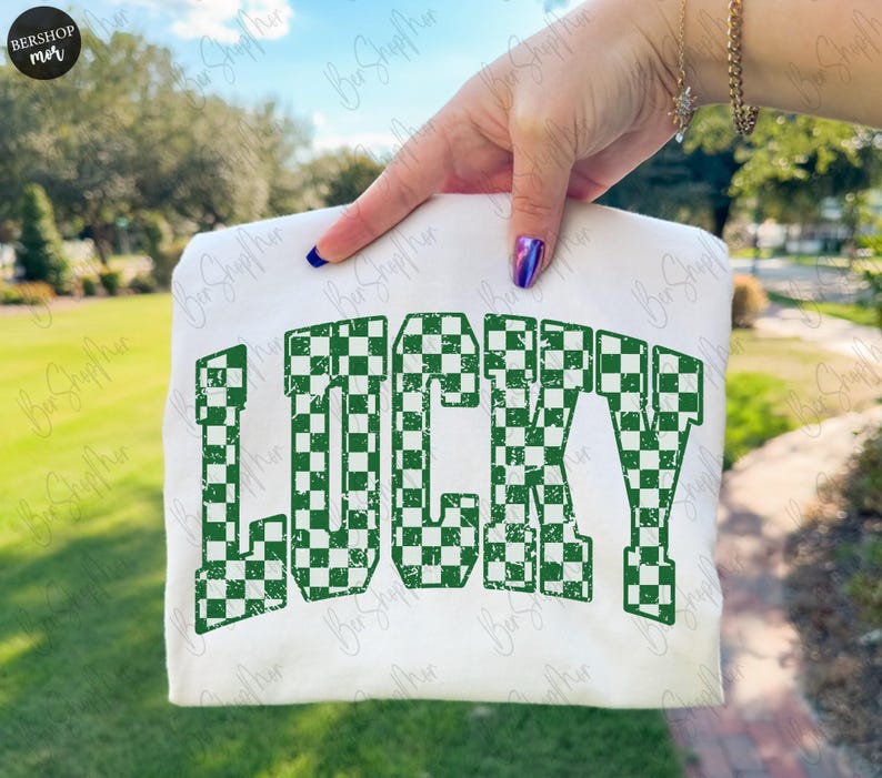 May include: A white t-shirt with the word "LUCKY" printed in large, green, checkered letters. The text has a distressed, vintage look. The shirt is held up against a blurred outdoor background.