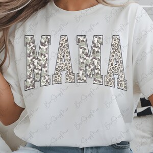 May include: White t-shirt with the word "MAMA" in large, block letters. The letters have a camouflage and leopard print design. The shirt is a casual style.