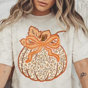 May include: Cream-colored t-shirt featuring a pumpkin design with a leopard print pattern. The pumpkin has an orange bow and leaves. The shirt has the text "BERSHOP mer" in the upper left corner.