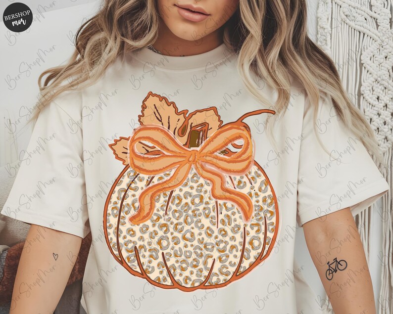 May include: Cream-colored t-shirt featuring a pumpkin design with a leopard print pattern. The pumpkin has an orange bow and leaves. The shirt has the text "BERSHOP mer" in the upper left corner.