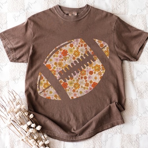 May include: Brown t-shirt featuring a floral football design. The football is filled with a pattern of orange, yellow, and pink flowers. The shirt has short sleeves and a crew neck. The shirt is a relaxed fit.