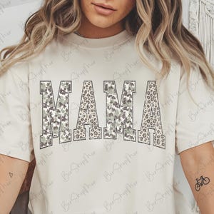 May include: Cream-colored t-shirt with the word "MAMA" in large, block letters. The letters have a camouflage and leopard print design. The shirt has short sleeves and a relaxed fit.