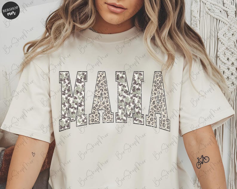 May include: Cream-colored t-shirt with the word "MAMA" in large, block letters. The letters have a camouflage and leopard print design. The shirt has short sleeves and a relaxed fit.