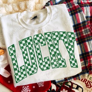 May include: A white sweatshirt with the word "LUCKY" in large, green checkered letters. The sweatshirt is part of a holiday-themed display, with a red and black plaid shirt and Christmas decorations in the background.