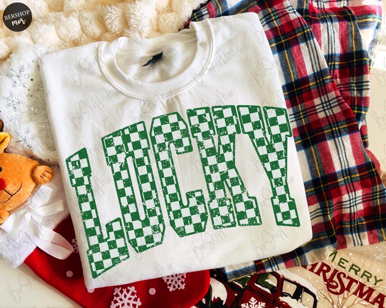 May include: A white sweatshirt with the word "LUCKY" in large, green checkered letters. The sweatshirt is part of a holiday-themed display, with a red and black plaid shirt and Christmas decorations in the background.
