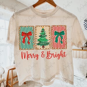 May include: Cream-colored t-shirt featuring a festive design. The design includes three rectangular panels with a red bow on green stripes, a Christmas tree, and a green bow on red stripes. The words "Merry & Bright" are written in red script.