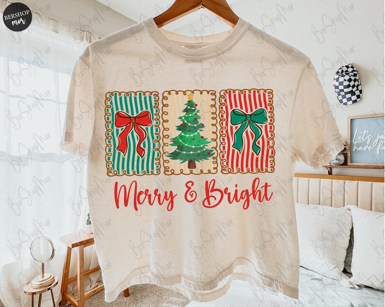 May include: Cream-colored t-shirt featuring a festive design. The design includes three rectangular panels with a red bow on green stripes, a Christmas tree, and a green bow on red stripes. The words "Merry & Bright" are written in red script.