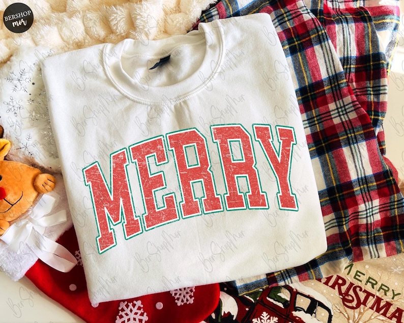 May include: A white crewneck sweatshirt featuring the word MERRY printed in a distressed red font with a green outline. The garment is displayed alongside red and green plaid fabric, a red stocking, and a plush reindeer toy.