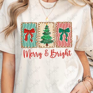 May include: Cream-colored t-shirt featuring a festive design. The design includes three rectangular panels with a red bow, a Christmas tree, and a green bow. The words "Merry & Bright" are written in red script below the panels.