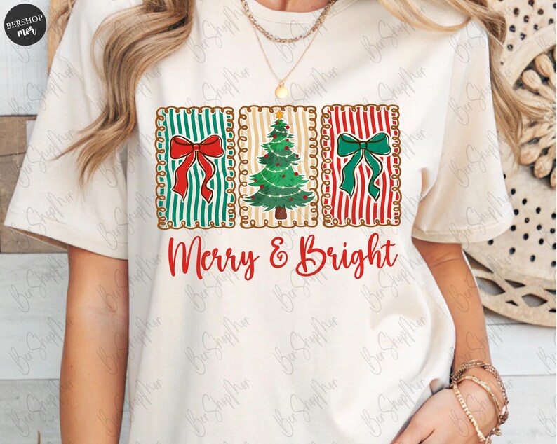 May include: Cream-colored t-shirt featuring a festive design. The design includes three rectangular panels with a red bow, a Christmas tree, and a green bow. The words "Merry & Bright" are written in red script below the panels.