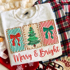 May include: White sweatshirt with a festive design featuring three rectangular panels. The panels display a Christmas tree and bows, with the text "Merry & Bright" in red script. The design is set against a backdrop of plaid fabric and Christmas stockings.