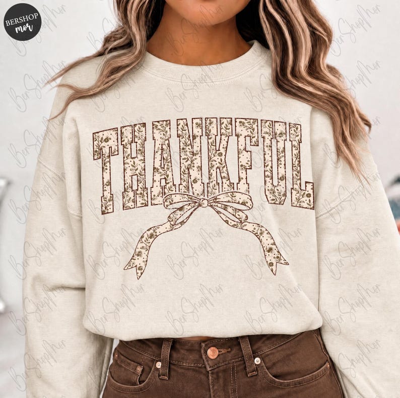 May include: Cream-colored sweatshirt with the word "THANKFUL" in a brown floral print. A matching bow is below the word. The sweatshirt has long sleeves and a crew neck. The model is wearing brown pants.