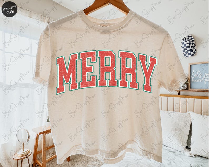 May include: A cream-colored short-sleeve t-shirt featuring the word MERRY printed in a large, arched, distressed red font with a dark green outline. The shirt is displayed on a wooden hanger against a light-colored interior background.