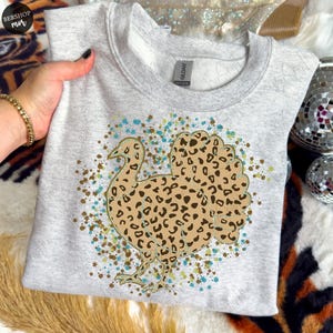 May include: A light gray sweatshirt featuring a turkey design with a leopard print pattern. The turkey is outlined in gold and surrounded by teal and brown speckles. The sweatshirt is a crew neck style.