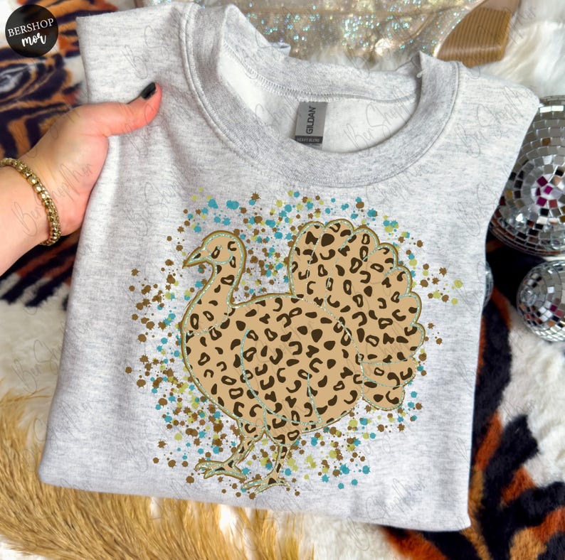 May include: A light gray sweatshirt featuring a turkey design with a leopard print pattern. The turkey is outlined in gold and surrounded by teal and brown speckles. The sweatshirt is a crew neck style.