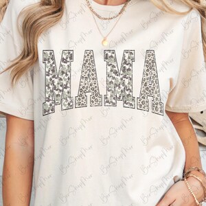 May include: Cream-colored t-shirt with the word "MAMA" in large, bold letters across the front. The letters have a camouflage and leopard print design. The shirt has short sleeves and a crew neck.
