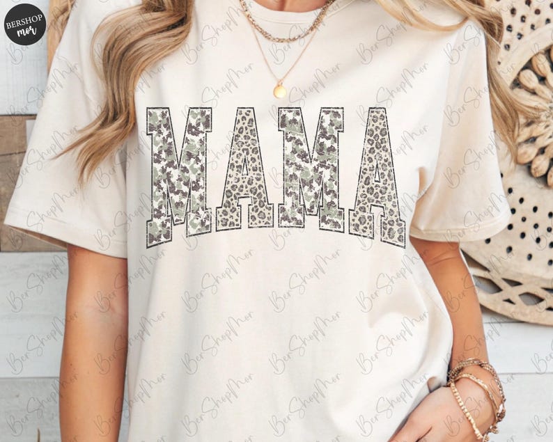 May include: Cream-colored t-shirt with the word "MAMA" in large, bold letters across the front. The letters have a camouflage and leopard print design. The shirt has short sleeves and a crew neck.