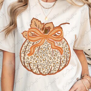 May include: Cream-colored t-shirt featuring a pumpkin design. The pumpkin has a leopard print pattern, an orange bow, and a stem with leaves. The design is centered on the shirt, creating a fall-themed aesthetic.
