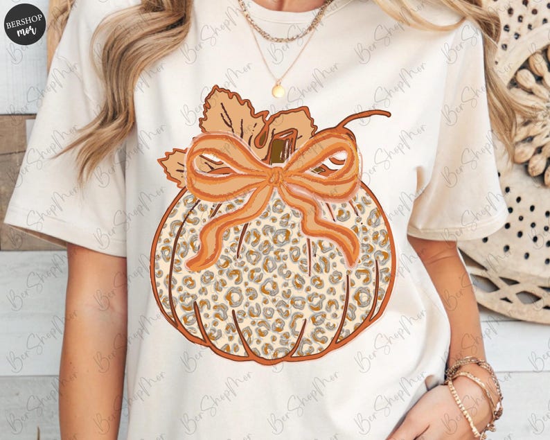 May include: Cream-colored t-shirt featuring a pumpkin design. The pumpkin has a leopard print pattern, an orange bow, and a stem with leaves. The design is centered on the shirt, creating a fall-themed aesthetic.