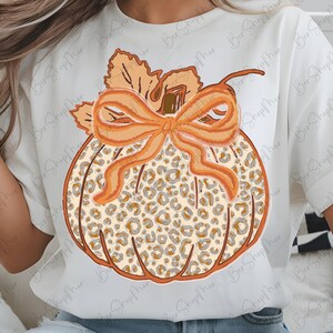 May include: White t-shirt featuring a pumpkin design with a leopard print pattern. The pumpkin has an orange bow and stem, with leaves at the top. The design is in shades of orange, brown, and beige, perfect for fall.