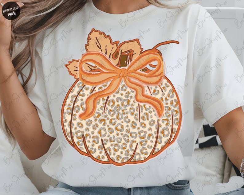 May include: White t-shirt featuring a pumpkin design with a leopard print pattern. The pumpkin has an orange bow and stem, with leaves at the top. The design is in shades of orange, brown, and beige, perfect for fall.