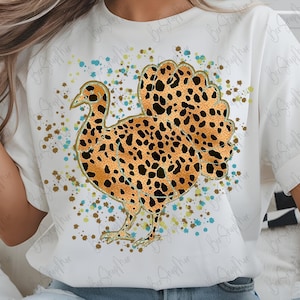 May include: White t-shirt featuring a turkey design with a leopard print pattern. The turkey is outlined in gold and surrounded by colorful paint splatters. The shirt has short sleeves and a crew neck.